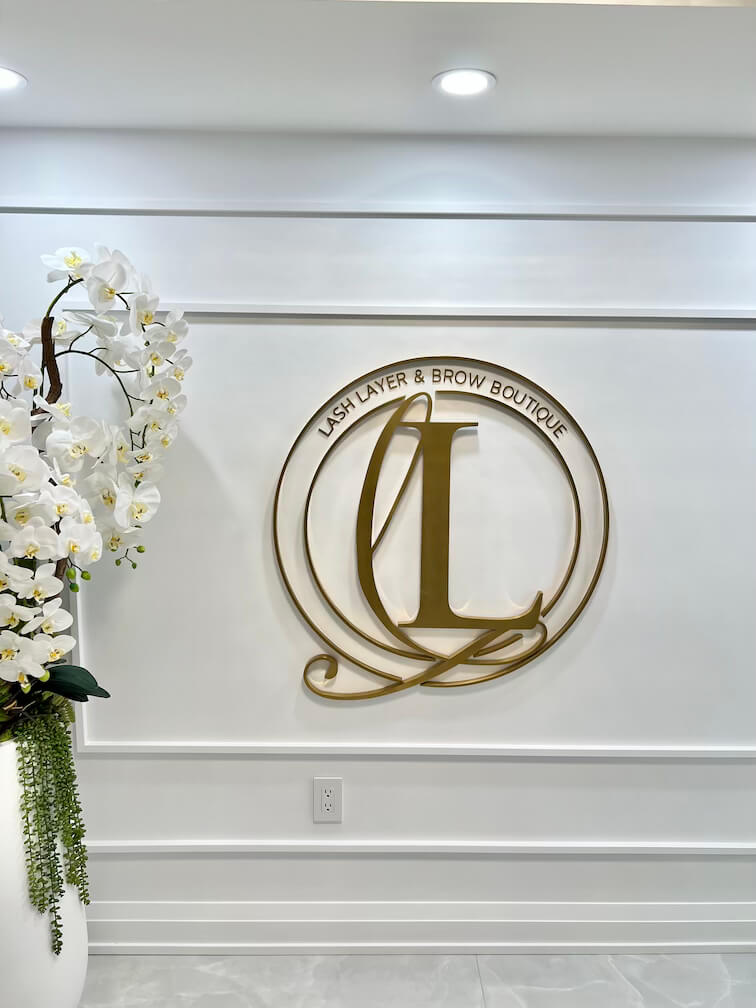 Chic entrance of Lash Layer & Brow Boutique in Pickering, Ontario – featuring a gold salon logo, modern lighting, and elegant white orchid décor.