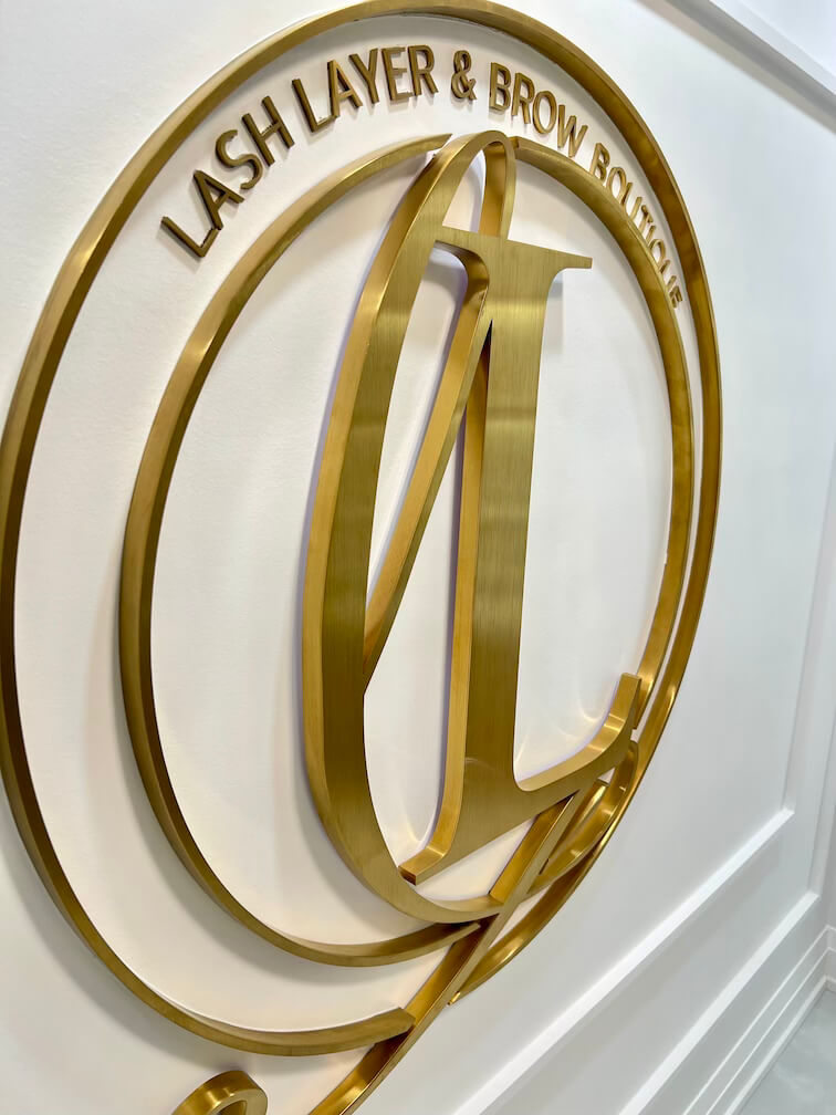 Close-up of the gold Lash Layer & Brow Boutique logo mounted on a white paneled wall in Pickering, Ontario.